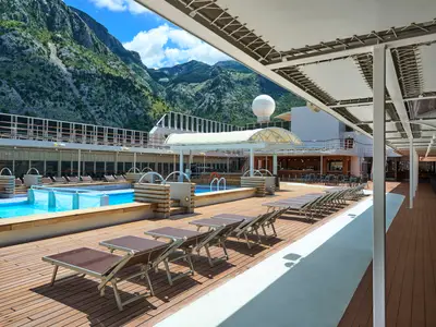 Blick auf das Pooldeck der MSC Armonia mit Pools, Liegestühlen, Bars und einem großen weißen Globus auf dem Oberdeck. Im Hintergrund erheben sich grüne, bewaldete Berge unter einem blauen Himmel mit weißen Wolken. Es ist ein sonniger Tag.