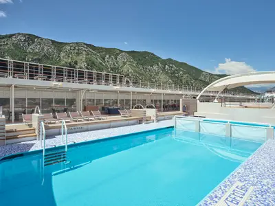 Blick auf den Poolbereich der MSC Armonia mit türkisfarbenem Wasser, umgeben von blau-weiß gefliesten Kanten und Holzterrassen. Im Hintergrund sind Liegestühle, Restaurants und grüne Berge unter einem klaren blauen Himmel zu sehen.