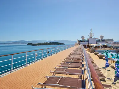 Blick über das Deck der MSC Armonia mit Reihen von braunen Liegestühlen. Im Hintergrund sind Pools mit blauen und grünen dekorativen Säulen und die Schiffsaufbauten zu sehen. Links erstreckt sich das weite blaue Meer mit einer bewaldeten Insel und einer Bergkette am Horizont unter klarem Himmel.