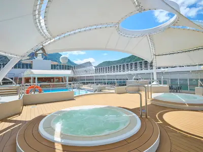 Blick auf das obere Deck der MSC Armonia mit drei runden Whirlpools und einem rechteckigen Pool. Links befindet sich eine Poolbar. Im Hintergrund erheben sich grüne Berge unter blauem Himmel mit einigen Wolken. Weiße Sonnensegel überspannen Teile des Decks.