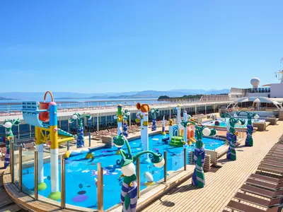 Blick von oben auf den lebhaften Doremi Wasserpark auf dem Deck der MSC Armonia. Bunte Spielstrukturen, Wasserrutschen und Spritzelemente sind in einem flachen Pool auf blauem Untergrund angeordnet. Im Hintergrund erstreckt sich eine Reihe von Liegestühlen entlang des Decks, während das Meer und eine bergige Küstenlinie unter einem klaren blauen Himmel zu sehen sind.
