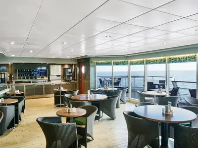 Blick in den Buffetbereich La Brasserie an Bord der MSC Armonia mit Holztischen und geflochtenen Stühlen. Große Fensterfront mit weitem Meerblick. Im Hintergrund eine Theke mit Kaffee- und Getränkeautomaten. Der Raum ist hell und ohne Personen.