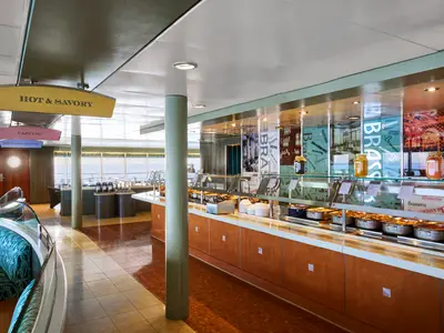 Blick in das helle Buffetrestaurant 'La Brasserie' an Bord der MSC Armonia mit warmer Beleuchtung und Blick auf das Meer. Links ein Sitzbereich, rechts das lange Buffet mit verschiedenen warmen Speisen, Tellern und Besteck. Deckenhänger weisen auf 'Hot & Savory', 'Carving' und 'Beverage Station' hin.