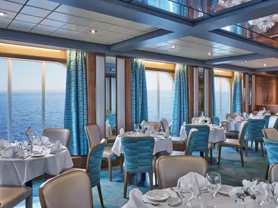 Elegantes Restaurant "La Pergola" an Bord der MSC Armonia. Gedeckte Tische mit Meerblick durch große Fenster. Blaue und cremefarbene Stühle, weiße Tischdecken und Holzvertäfelung.