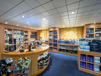 Blick in den gut beleuchteten Bordshop an Bord der MSC Armonia, gefüllt mit Souvenirs, Markenkleidung, Elektronik und Snacks. Blaue Teppichböden und goldene Vorhänge rahmen die Auslagen ein.