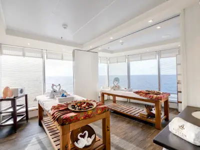Helle Spa-Massagekabine an Bord der MSC Armonia. Zwei Massageliegen mit weissen Handtüchern und gefalteten Schwänen. Grosse Fenster mit Meerblick. Salzkristalllampe und Potpourri sind sichtbar.