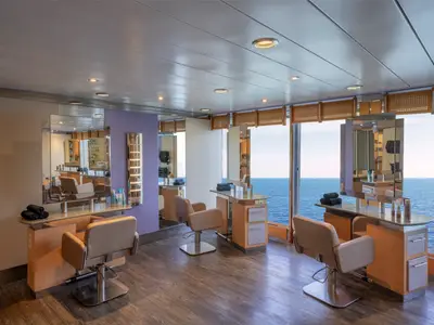 Moderner Hair Salon im MSC Aurea Spa der MSC Armonia. Mehrere Friseurstühle, Spiegel und große Fenster mit Blick auf das weite Meer. Helle Holzmöbel und beige Stühle prägen den Raum.