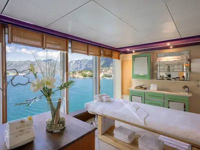 Der Spa-Behandlungsraum der MSC Armonia mit Massageliege und Produkten. Große Fenster bieten einen weiten Blick auf eine Bucht mit Bergen und Ortschaft. Der Raum ist hell und entspannend.