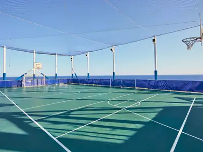 Ein leerer Multifunktions-Sportplatz mit grünem Boden, weißen Linien, Basketballkörben und Fußballtoren an Deck der MSC Armonia. Ein Schutznetz spannt sich darüber zum blauen Meer und Himmel.
