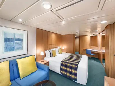 Eine helle Suite an Bord der MSC Armonia mit einem Doppelbett, einem blauen Sofa mit gelben Kissen, einem Schreibtisch mit blauem Stuhl und einem Wandspiegel. Die Wände sind holzgetäfelt, der Boden ist mit blaugrünem Teppich ausgelegt.