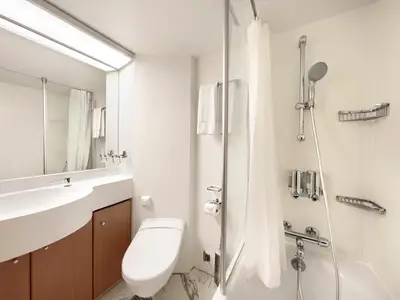 Helles Badezimmer an Bord der MSC Armonia mit einem modernen Waschbecken, einer Toilette und einem Duschbereich mit Duschvorhang und integrierten Seifenspendern. Ein Spiegel hängt über dem Waschbecken, und Handtücher sind ordentlich auf einem Handtuchhalter drapiert.