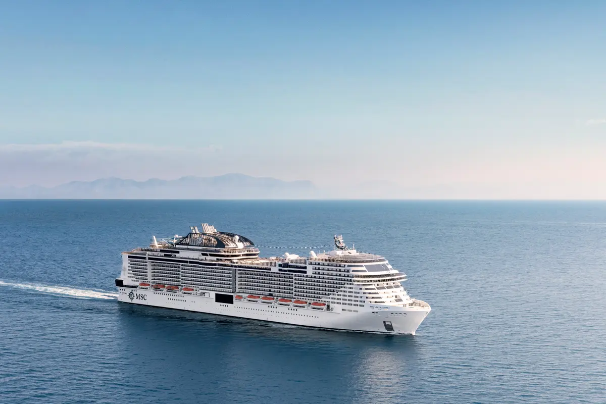 Die MSC Bellissima, ein modernes Kreuzfahrtschiff, fährt auf blauem Meer. Berge und blauer Himmel am Horizont. Ein weißer Kielwasserstreifen ist hinter dem Schiff zu sehen.
