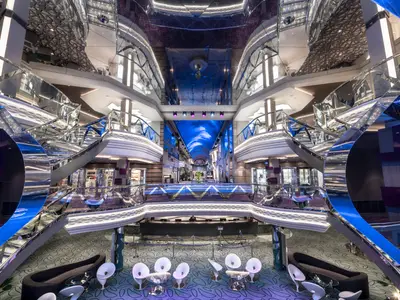 Ein Weitwinkelbild des eleganten Infinity Atriums der MSC Bellissima. Symmetrische Treppen mit Swarovski-Kristallen flankieren einen digitalen Himmel. Im Vordergrund eine Rezeption und Sitzbereiche.
