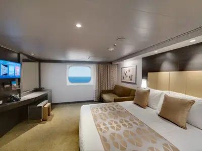 Eine helle und moderne Aussenkabine an Bord der MSC Bellissima. Im Vordergrund ein grosses Doppelbett, im Hintergrund ein Sofa und ein Fenster mit Blick auf das Meer. Links ein Schreibtisch mit TV.