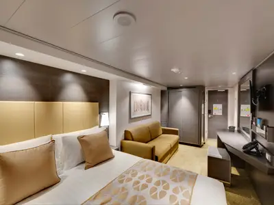 Deluxe Balkonkabine an Bord der MSC Bellissima. Ein Doppelbett, Sofa und ein Schreibtisch mit TV und Spiegel sind sichtbar. Moderne, warme Einrichtung mit indirekter Beleuchtung.