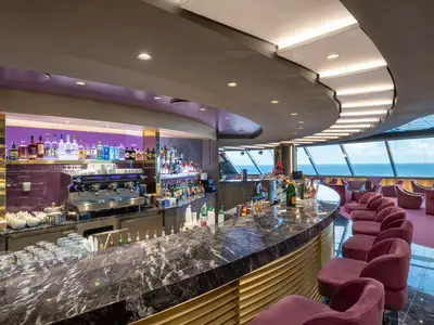 Eine elegante Bar-Lounge an Bord der MSC Bellissima. Eine lange, dunkle Marmorbar mit vielen Flaschen und einem Kaffeeautomaten ist zu sehen, davor stehen violette Barhocker. Große Fensterfronten bieten einen weiten Blick auf das offene Meer.