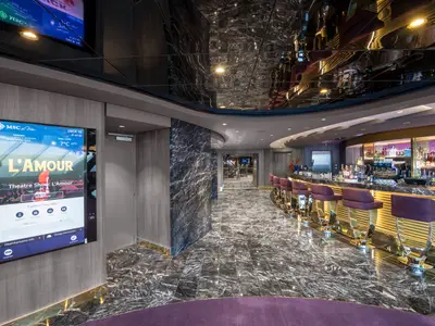 Die stilvolle Top Sail Lounge an Bord der MSC Bellissima zeigt eine elegante Bar mit violetten Hockern und goldenen Akzenten. Ein interaktiver Bildschirm links zeigt Bordinformationen.