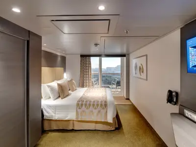 Geräumige Balkonkabine an Bord der MSC Bellissima mit einem grossen Doppelbett, moderner Einrichtung, einem Wandfernseher und einem Balkon mit Blick auf den Hafen und eine Stadt-Skyline.