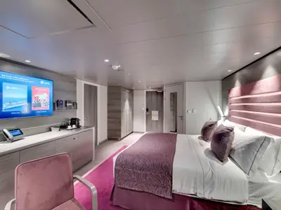 Innenansicht einer modernen Suite an Bord der MSC Bellissima. Das Zimmer verfügt über ein Doppelbett mit rosafarbener Kopflehne und einen großen Fernseher.