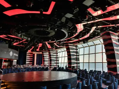 Weitwinkelaufnahme der futuristischen Carousel Lounge der MSC Bellissima. Eine runde Bühne, viele dunkelblaue Sitze und eine auffällige Deckenkonstruktion mit roten Lichtern prägen den Raum. Große Fenster bieten Meerblick.