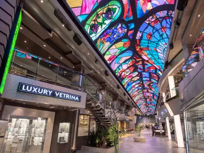 Die helle Galleria Bellissima an Bord der MSC Bellissima, einer von Geschäften gesäumten Promenade, wird von einem farbenprächtigen LED-Himmel überspannt. Links ein Schild 'LUXURY VETRINA'.