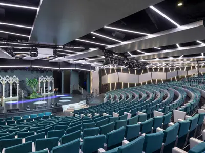 Weitwinkelansicht des eleganten London Theatres an Bord der MSC Bellissima. Die Buehne ist mit gotischen Boegen und einem Baum dekoriert. Der Zuschauerraum ist mit zahlreichen Reihen von teal-farbenen Sitzen bestueckt. Die Decke und Waende sind mit weissen LED-Lichtleisten beleuchtet, die eine dynamische Atmosphaere schaffen.
