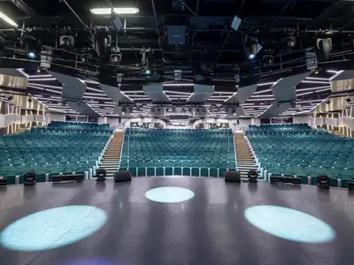Blick auf das leere London Theatre an Bord der MSC Bellissima. Blaue Sitzreihen steigen zu beiden Seiten der Mittelgänge an, während auf der Bühne drei beleuchtete Kreise Akzente setzen. Die Decke ist mit Lichtstreifen modern gestaltet.