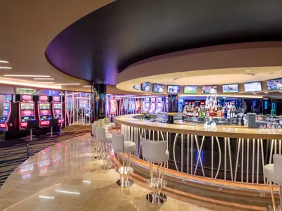 Elegantes Imperial Casino an Bord der MSC Bellissima mit zentraler, beleuchteter Bar, Spirituosen und Bildschirmen. Links moderne Spielautomaten.