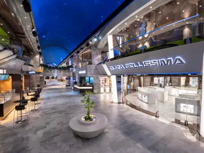 Die moderne, mehrstöckige Promenade Plaza Bellissima an Bord der MSC Bellissima. Überdacht von einem Sternenhimmel, bietet sie Geschäfte, Bars und Sitzgelegenheiten in hellem Ambiente.