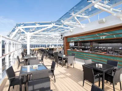 Grosszügiger Aussen-Buffetbereich an Deck der MSC Bellissima mit Tischen, Stühlen und einer langen Buffetheke unter einem Glasdach. Das Meer ist im Hintergrund sichtbar.