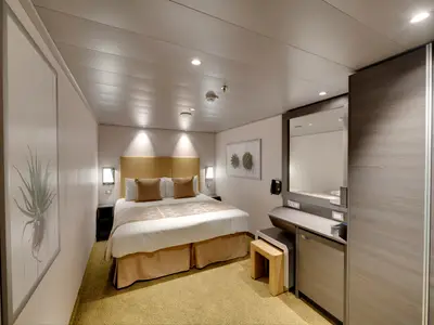 Moderne Innenkabine an Bord der MSC Bellissima mit einem komfortablen Doppelbett, Nachttischen und einer eleganten Schminkecke. Die Wände sind hell, der Teppich goldbraun. Pflanzenbilder schmücken die Wände.