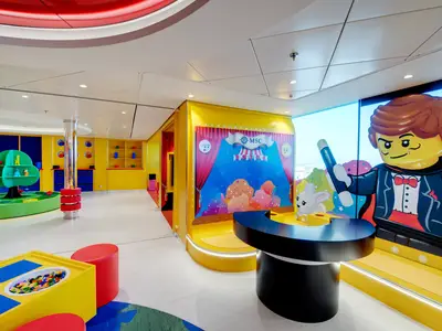 Der farbenfrohe Junior Club an Bord der MSC Bellissima zeigt einen Lego-Spielbereich mit einem riesigen Lego-Magier, einem Hasen und einem Trickkasten. Im Hintergrund eine kleine Bühne mit Vorhang und ein Spielbaum für Kleinkinder.