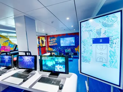 Der lebhafte "Magic Game Room" an Bord der MSC Bellissima. Mehrere Computertische und ein großer interaktiver Bildschirm, umgeben von bunten Legofiguren und Weltraumthemen.