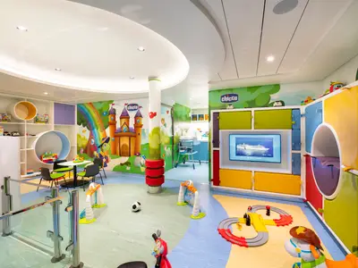 Heller, farbenfroher Baby- und Kleinkindspielbereich an Bord der MSC Bellissima. Bunte Wandmalereien, diverse Spielbereiche, kleine Tische und ein Monitor mit Kreuzfahrtschiff sind zu sehen. Keine Personen.