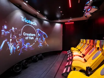 XD-Kino an Bord der MSC Bellissima. Ein großer, gebogener Bildschirm zeigt 'Triotech Creators of fun' und animierte Kreaturen. Gelbe Sitze mit Sicherheitsgurten stehen bereit. Projektoren hängen von der Decke.