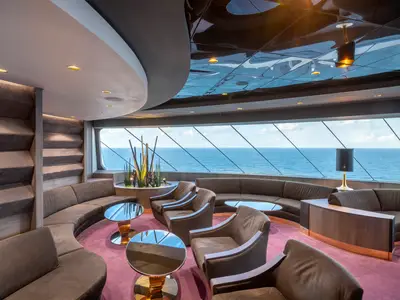 Elegante Top Sail Lounge an Bord der MSC Bellissima. Große Fensterfront mit Meerblick. Samtige Sofas und Sessel, Couchtische und Pflanzendeko. Indirekte Beleuchtung.
