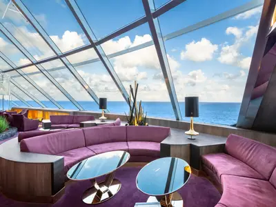 Die elegante Top Sail Lounge an Bord der MSC Bellissima mit violetten Sofas und modernen Tischen. Grosse Glasfenster bieten einen weiten Blick auf das blaue Meer und den Himmel.