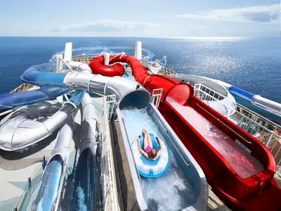 Ein Mädchen auf einem blauen Reifen rutscht die rote Wasserrutsche des Polar Aquaparks auf dem oberen Deck der MSC Bellissima hinunter. Weitere bunte Rutschen und das glitzernde blaue Meer sind im Hintergrund zu sehen.