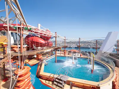 MSC Bellissima, Arizona Aquapark: Rote Wasserrutschen, Pool, Wasserspiele und Hochseilgarten auf dem Sonnendeck. Hafenstadt im Hintergrund.