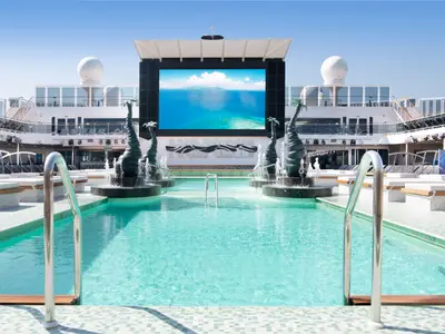 MSC Bellissima, Atmosphere Pool: Türkisfarbenes Wasser, Liegeflächen, Palmen-Brunnen und grosse LED-Leinwand mit tropischem Motiv. Blauer Himmel, helles Tageslicht.
