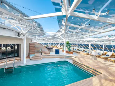 Blick auf den grosszügigen, überdachten Poolbereich mit türkisem Pool, Holzdeck, vielen Liegen und Blick auf das weite Meer an Bord der MSC Bellissima.