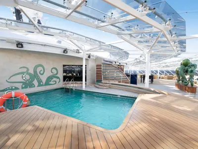 Der Poolbereich des MSC Yacht Club an Bord der MSC Bellissima. Türkisfarbener Pool, Holzdeck, Wandkunst mit Krakenmotiven, eine grosse grüne Oktopus-Skulptur und eine moderne Glasdachkonstruktion.