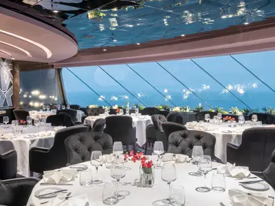 Innenansicht des eleganten MSC Yacht Club Restaurants an Bord der MSC Bellissima, mit Tischen, schwarzen Stühlen und großen Panoramafenstern zum Meer.