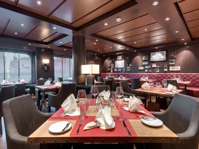 Blick in das stilvolle Steakhouse Butcher's Cut an Bord der MSC Bellissima mit gedeckten Tischen, bequemen Stühlen und einer langen roten Sitzbank. Die Wände sind mit Bildern dekoriert, während durch die Fenster Tageslicht hereinströmt.