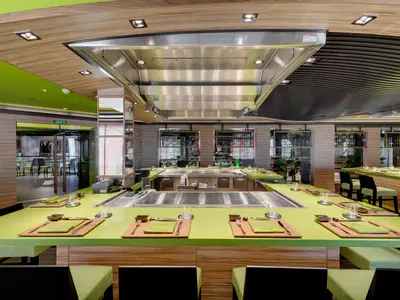 Das Kaito Teppanyaki Restaurant an Bord der MSC Bellissima. Es sind mehrere moderne Kochstationen mit hellgrünen Tischen, Gedecken und Holzelementen zu sehen. Keine Personen.