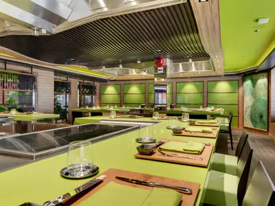 Restaurant an Bord der MSC Bellissima. Lange, hellgrüne Tische für Teppanyaki mit Kochstationen, gedeckt. Wände mit Holz und grünen Paneelen, dazu grüne Schirm-Kunstwerke. Modernes, elegantes Design.
