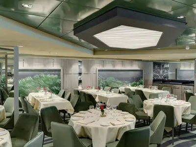 Blick in das elegante Posidonia Restaurant an Bord der MSC Bellissima. Zahlreiche Tische mit weissen Tischdecken, grüne Stühle und Wandgemälde prägen den Raum. Moderne Deckenbeleuchtung. Ohne Personen.