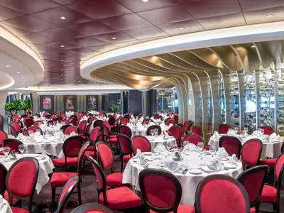 Hauptrestaurant Il Ciliegio an Bord der MSC Bellissima. Viele festlich gedeckte Tische mit weißen Tischdecken und roten Stühlen. Die moderne Architektur zeigt geschwungene Deckenelemente und ein großes Weinregal.