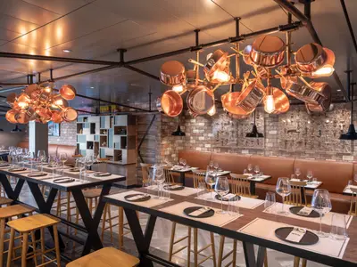 Innenansicht des Spezialitätenrestaurants Hola! Tapas Bar by Ramon Freixa an Bord der MSC Bellissima. Mit langen Holztischen, Lederbänken und einzigartigen Kupferpfannen-Deckenlampen.