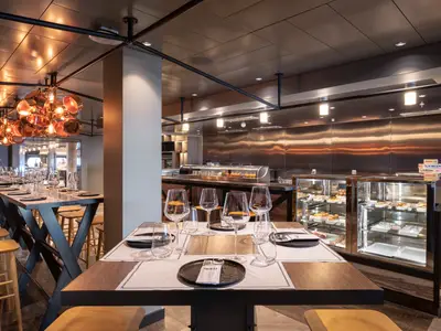 Innenansicht der Hola! Tapas Bar an Bord der MSC Bellissima. Elegante Tische mit Weingläsern sind bereit. Im Hintergrund ist eine Theke mit Speisen und eine Wand aus Edelstahl zu sehen.